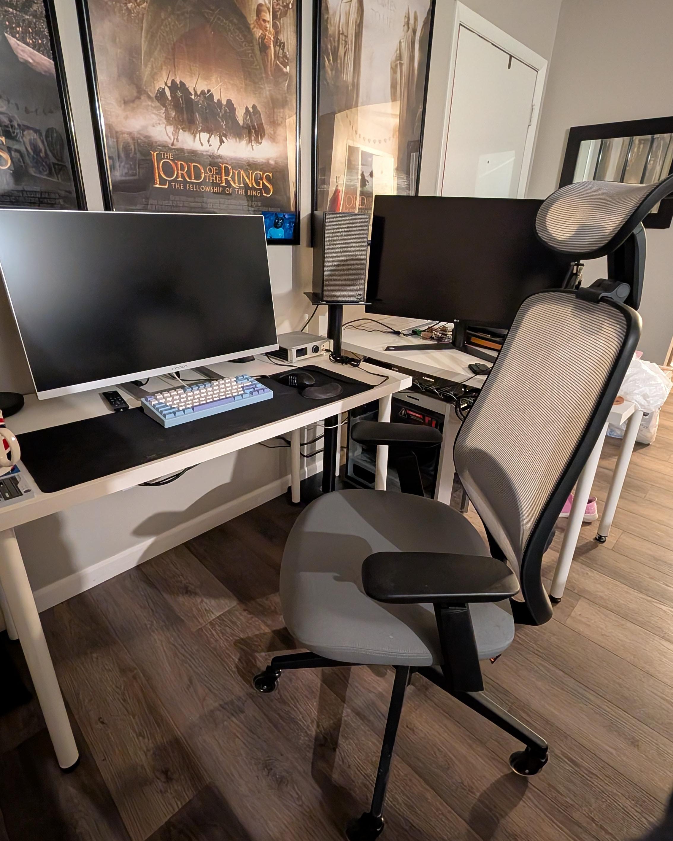 A modern gray and black mesh office chair in a home office setup, positioned in front of a white desk with dual computer monitors, wood flooring, and framed Lord of the Rings posters on the wall.
