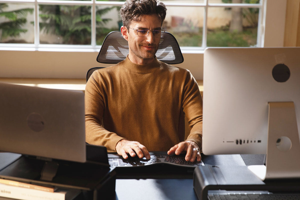 A man in a brown sweater is working at a desk with computers.