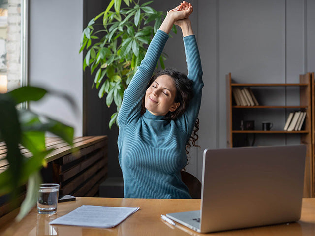 A person stretching their arms at a desk in a modern office with green plants and bookshelves in the background.