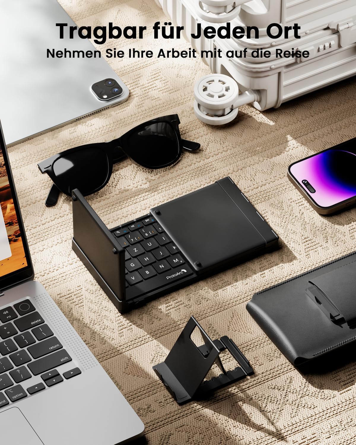 Black foldable Bluetooth keyboard with DE layout, open on a table with laptop, phone, stand, and sunglasses.