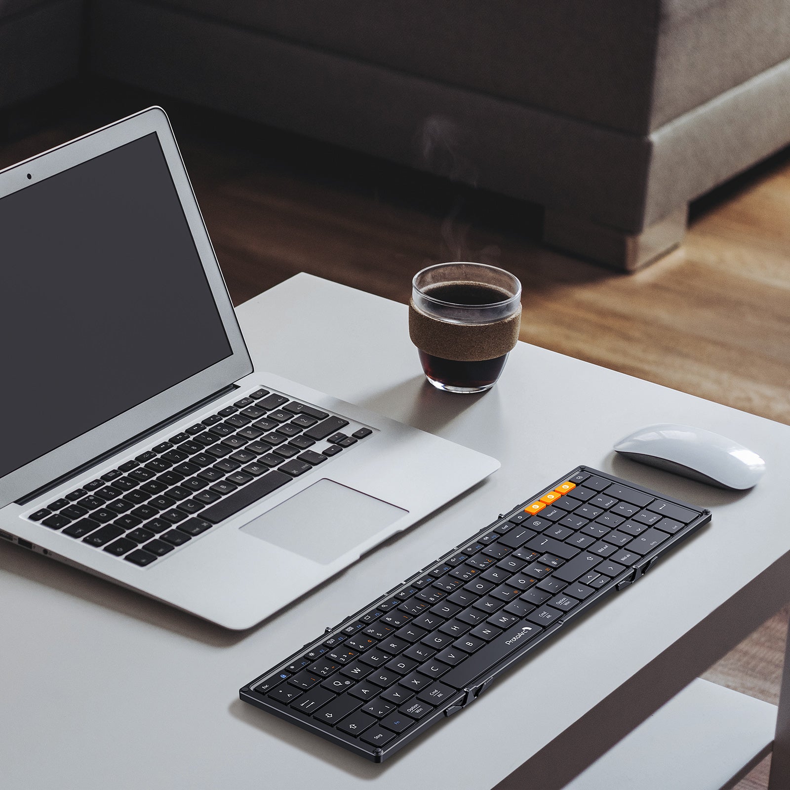 Black XK01 Tri-Fold Keyboard DE layout with orange keys next to a silver laptop and white mouse on a desk.