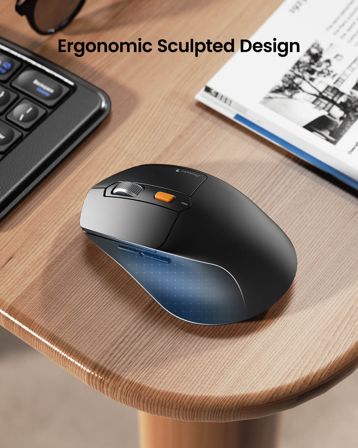 Ergonomic sculpted design black wireless mouse with orange button, placed on a wooden desk next to a keyboard and book.
